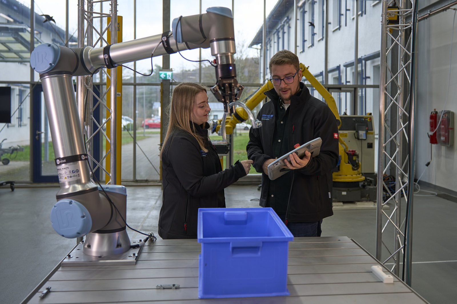 A woman and a man at a collaborative robot arm; the man holds a tablet, a blue box stands on the table.