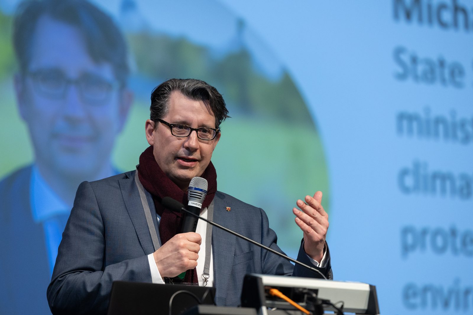 Ein Mann mit Brille und Schal spricht vor einer Projektion ins Mikrofon.