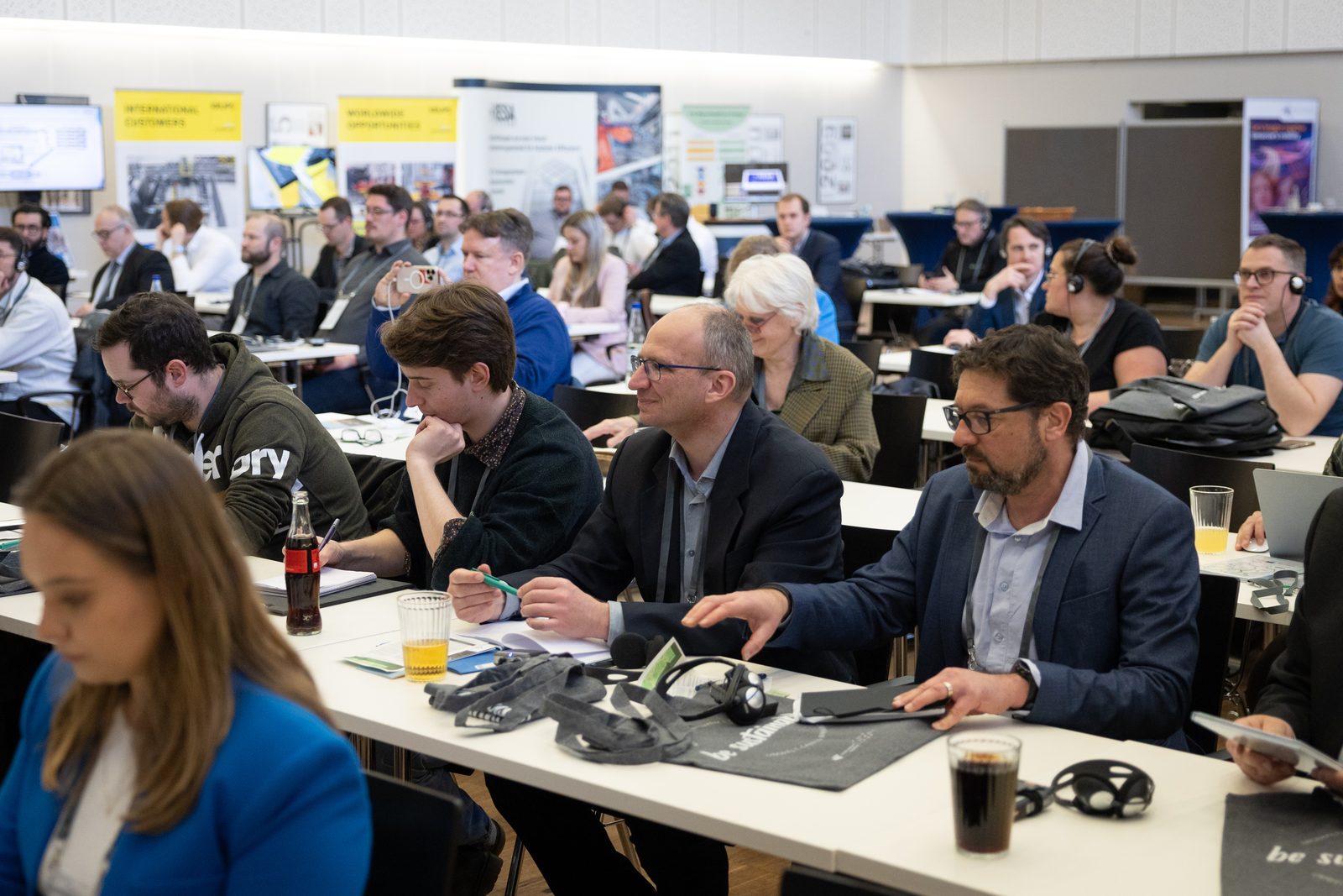 Teilnehmende an Tischen im Saal, einige mit Dolmetsch-Kopfhörern.
