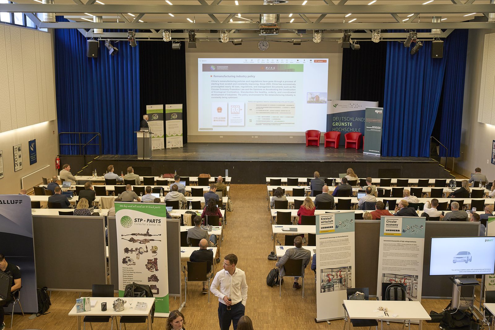 View over the full lecture hall with exhibition stands at the sides and a talk on stage.
