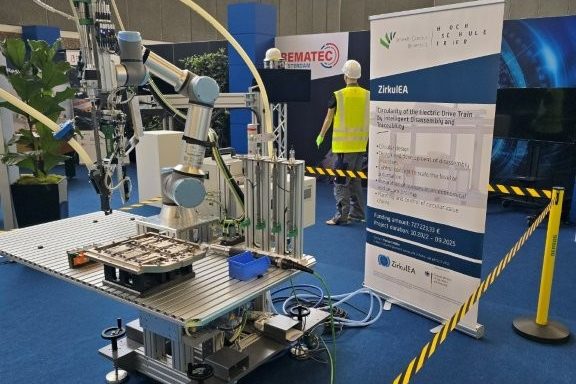 Eine Roboterzelle mit kollaborierendem Roboterarm und Werkzeugen neben einem Roll-up von ZirkulEA, mit gelb-schwarzem Band abgesperrt.