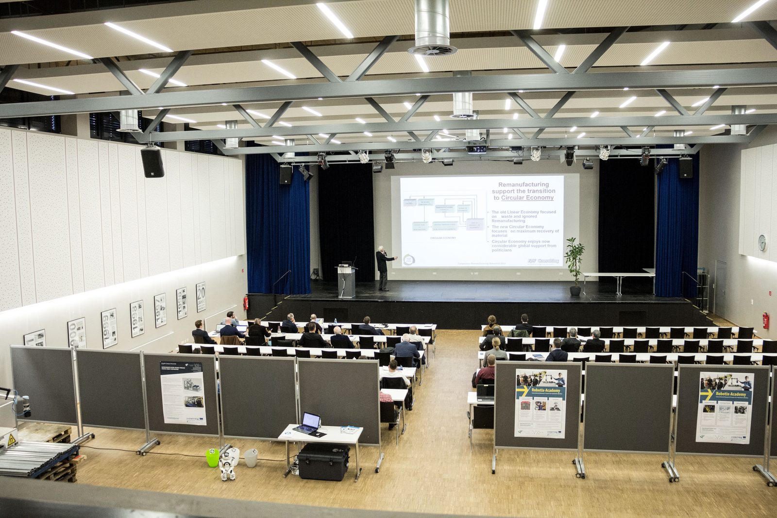 View over the lecture hall with exhibition stands and a talk on the screen.