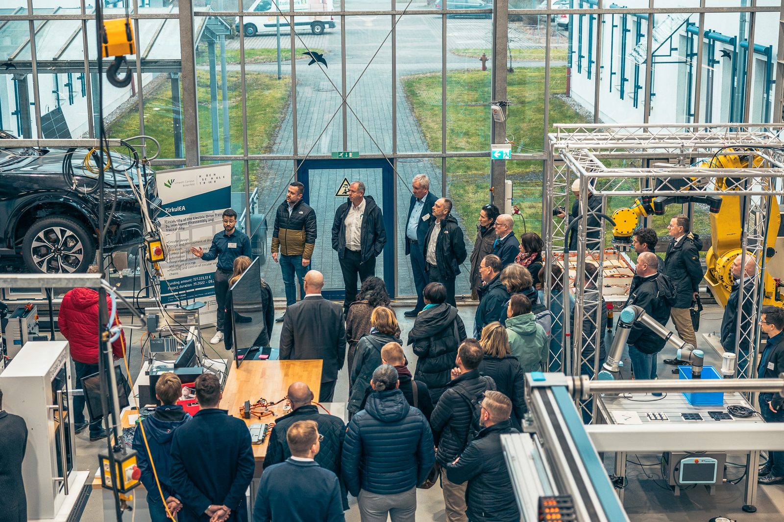 A large group of participants in a glass-roofed lab hall between robot cells.