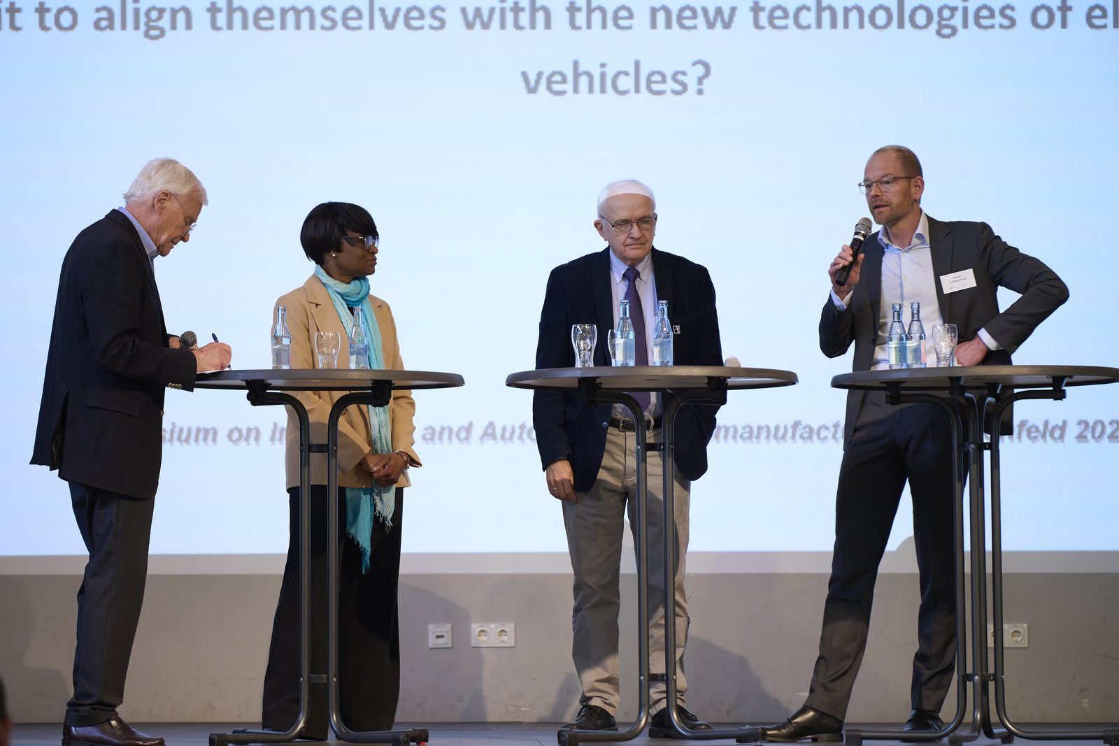 Four people at standing tables on a stage in front of a projection screen; one speaks into a microphone.